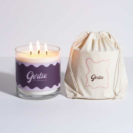 Lit gertie candle with violet label and a matching drawstring bag on a light gray background