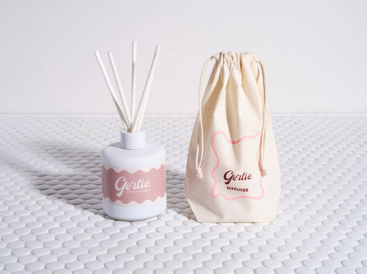A reed diffuser with white sticks in a white bottle that has 'Gertie' written on it, alongside a beige reusable cotton pouch with 'Gertie' in pink lettering.
