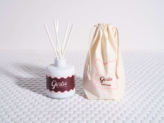 A Cherry Vanilla Reed Diffuser with a white vase, black and red label, and multiple reeds, alongside a beige reusable cotton pouch with red hearts and the brand name 'Gertie'.