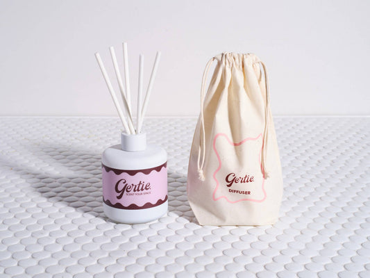 A reed diffuser with white sticks in a white bottle, labeled 'Gertie', next to a beige drawstring bag with the same brand on a textured white surface.