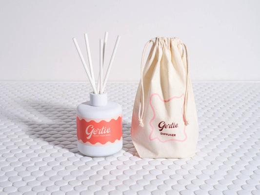 A white reed diffuser with a pink label and a set of white reeds next to a beige drawstring bag with the brand name 'Gertie' on it.