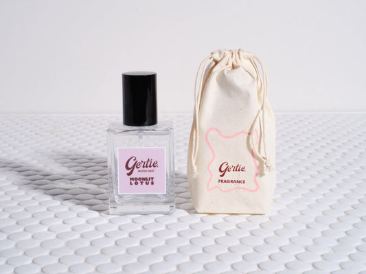 A bottle of 'Moonlit Lotus Room Mist' with a pink label and black cap, accompanied by a beige drawstring bag with pink text on it, displayed on a textured white surface.