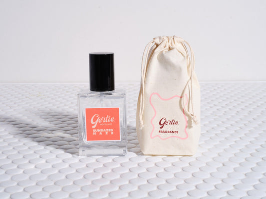A bottle of Sundaze Haze Room Mist with a black cap and a label featuring the product name and brand. Next to it is a beige drawstring bag with the brand logo.
